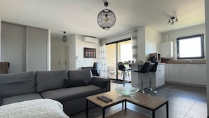 A modern living room with a grey sofa, a small coffee table, and a white throw blanket. There is a kitchen area with white cabinets and a window.