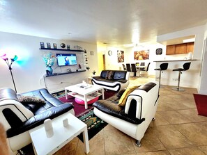 A living room with a sofa, coffee table, and TV. There is a dining area with chairs and a kitchen in the background.