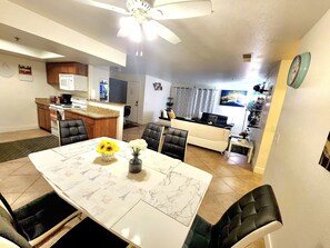 A dining area with a table set for four, a kitchen in the background, and a living area with a sofa and a TV.