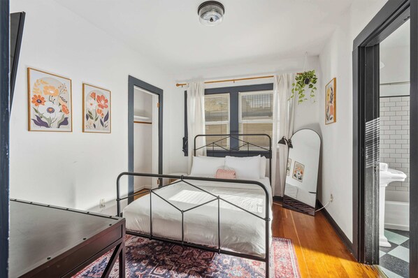 A bedroom with a metal bed frame, a bedside table, a patterned rug, a window with blinds, and a bathroom with a white toilet visible through an open door.