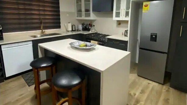 A modern kitchen with a central island, two bar stools, a refrigerator, and pendant lighting.