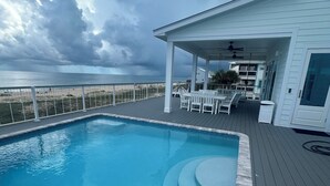 En swimmingpool med ett trädäck, en täckt uteplats med utemöbler och utsikt över stranden och havet.