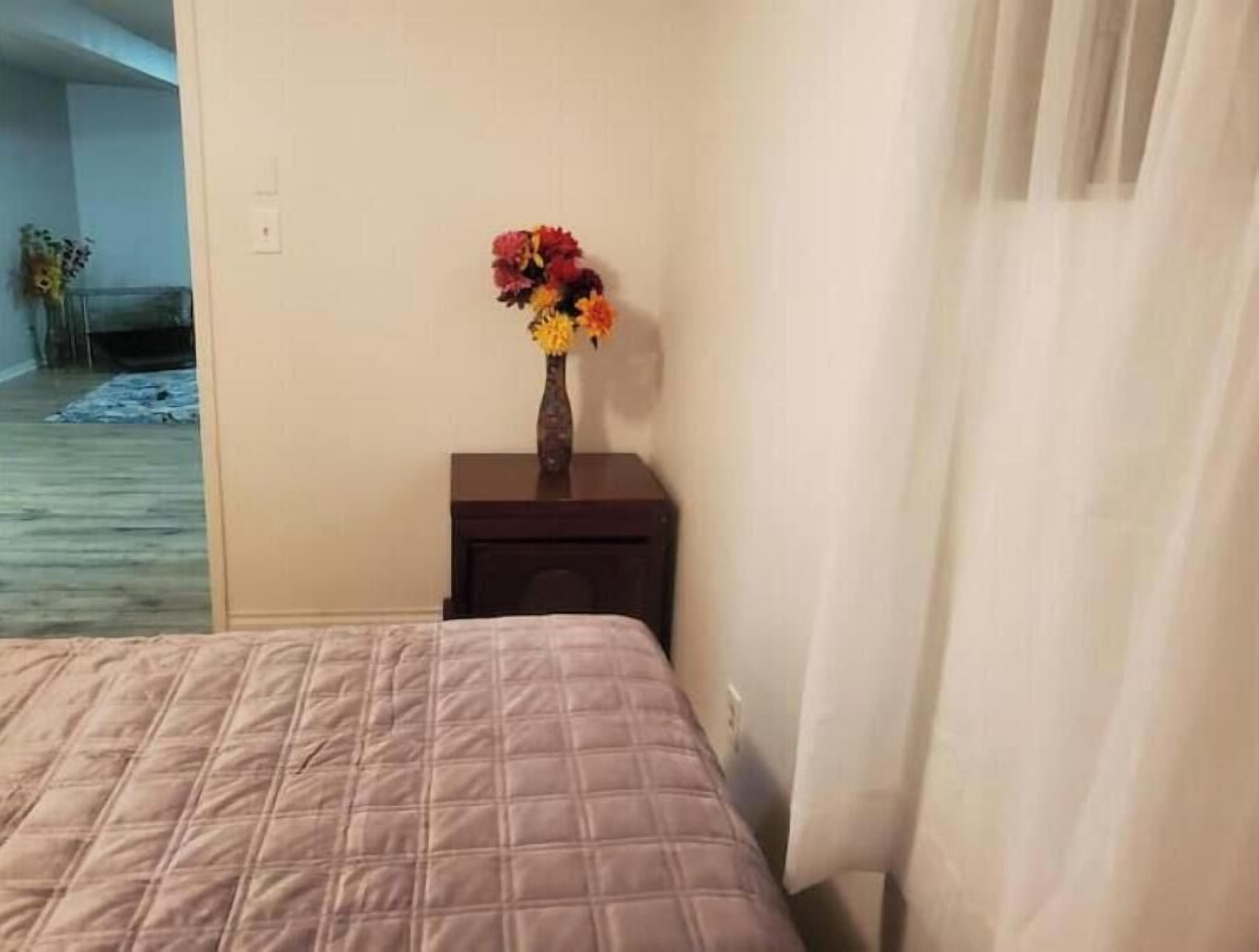 Quarto com uma cama, mesa de cabeceira com um vaso de flores e vista para um corredor.