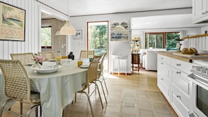 A spacious kitchen with white cabinetry, a dining area with wicker chairs and a striped tablecloth, and a view of greenery outside.