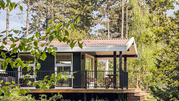 A modern cabin with a wooden deck, large windows, and a covered outdoor seating area surrounded by trees.