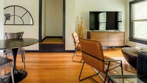 A modern living room with a wooden floor, a round dining table, leather chairs, a sofa, a TV on a wooden cabinet, and a large window with blinds. - Moab Vibes in PDX - Walk to Local Coffee & Food (Portland)