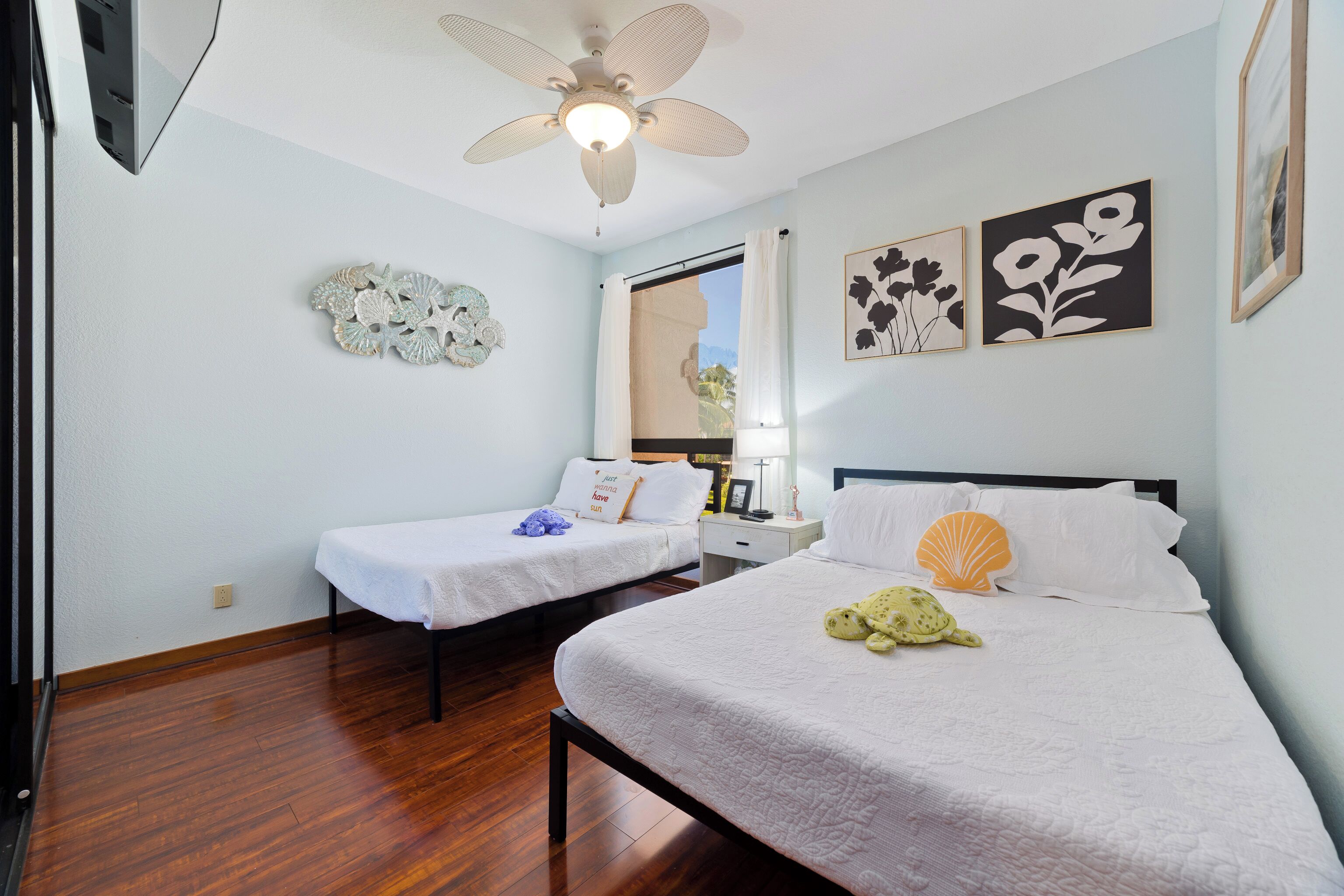 A hotel room with two beds, wooden flooring, a ceiling fan, and wall art.