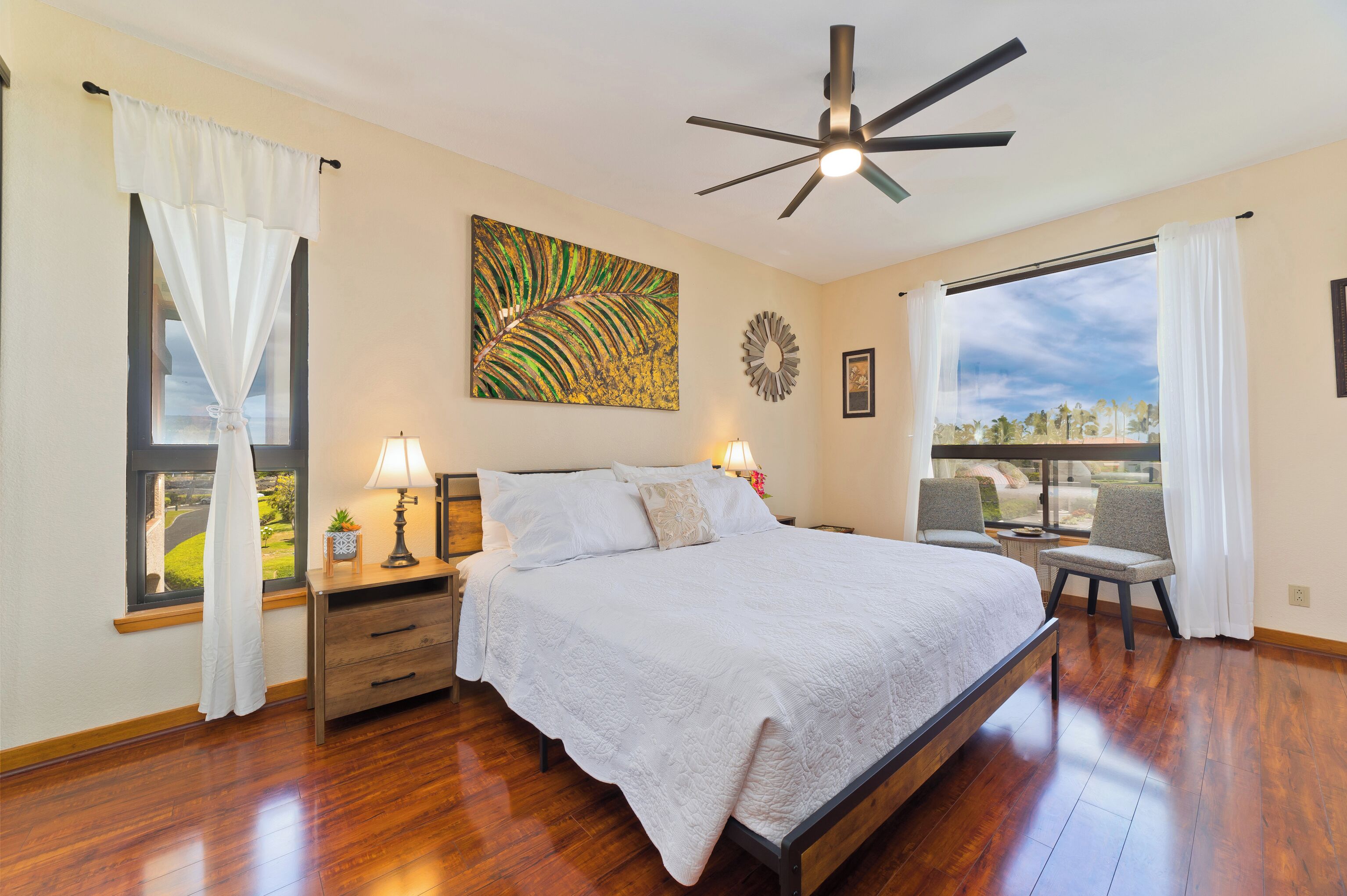 Une chambre avec un grand lit, un ventilateur de plafond, une fenêtre avec des rideaux, une table de chevet avec une lampe et une vue sur l’extérieur.