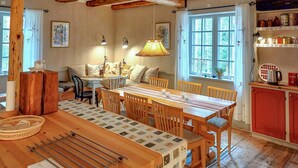 Een gezellige keuken met een houten eettafel, stoelen en een hanglamp. Er is een raam met gordijnen, een houten kast en een plank met keukenspullen.