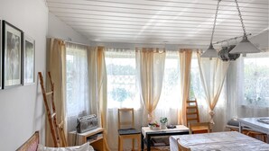 A room with a wooden ceiling, a table with a white tablecloth, a chair, a bed with a white pillow, and a radio on a shelf.