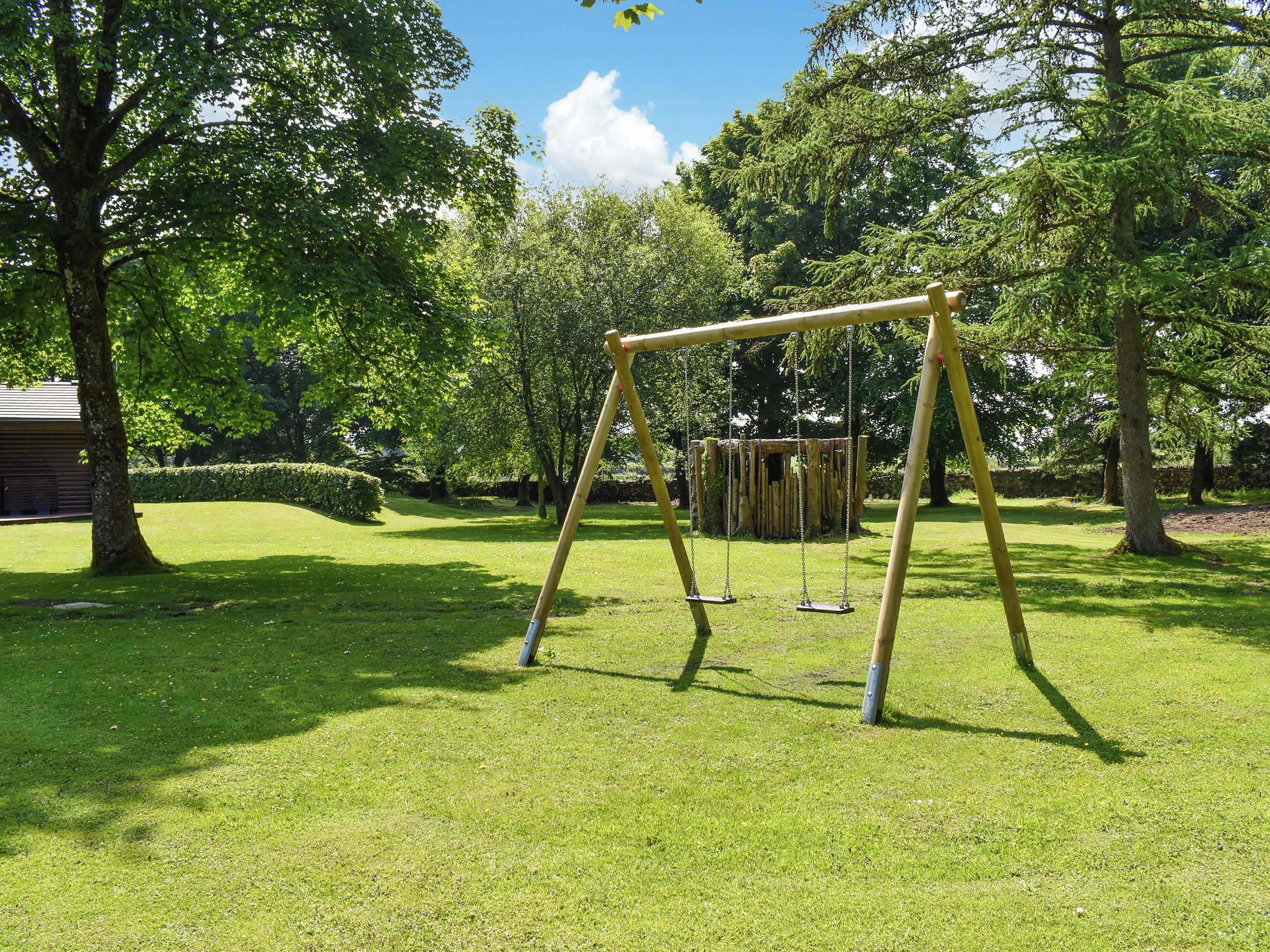 A swing set in a grassy park area with trees and a wooden structure in the background.