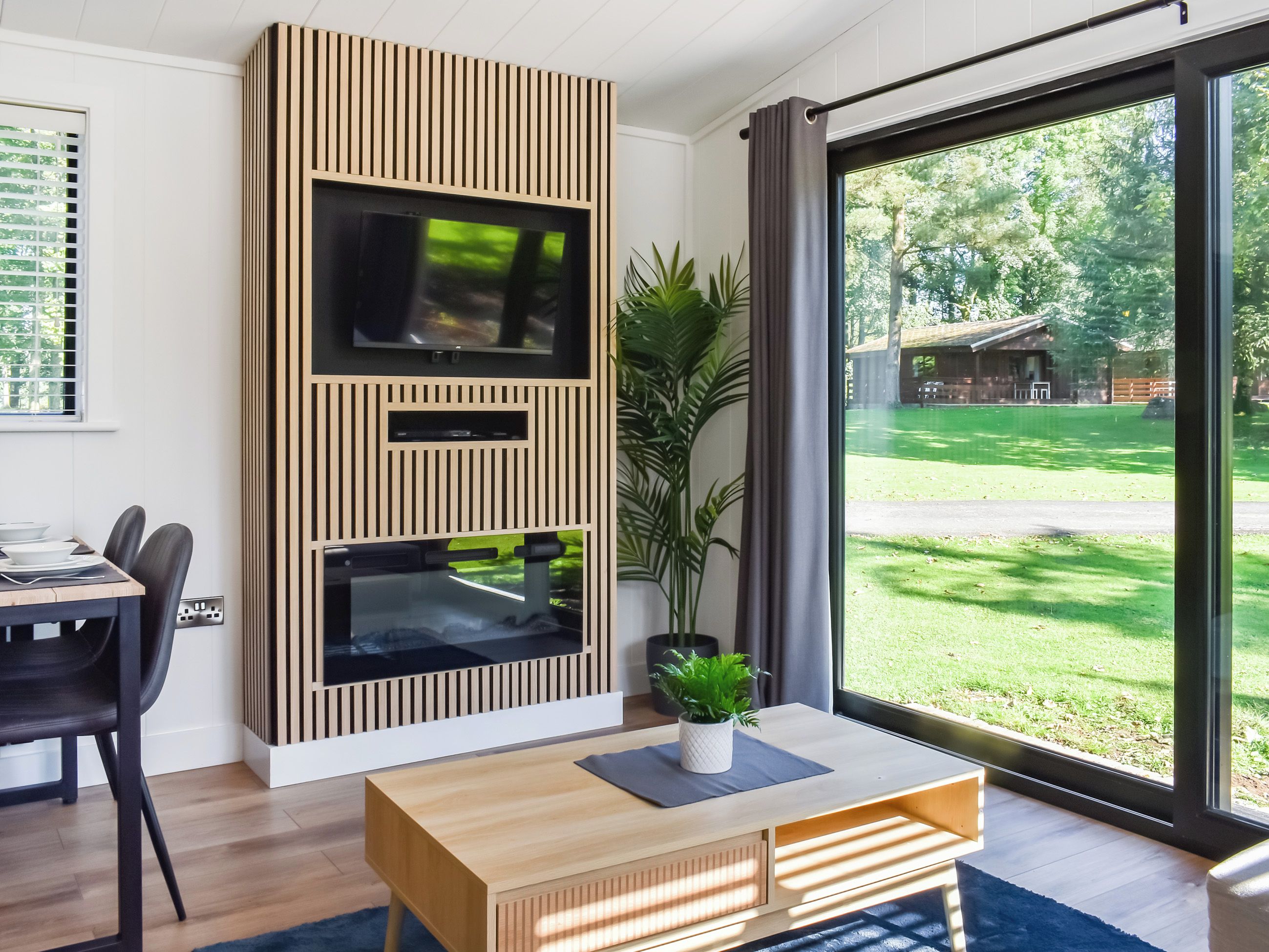 A modern living room with a TV, a dining area with chairs, a wooden coffee table, and a large window with a view of a green landscape.