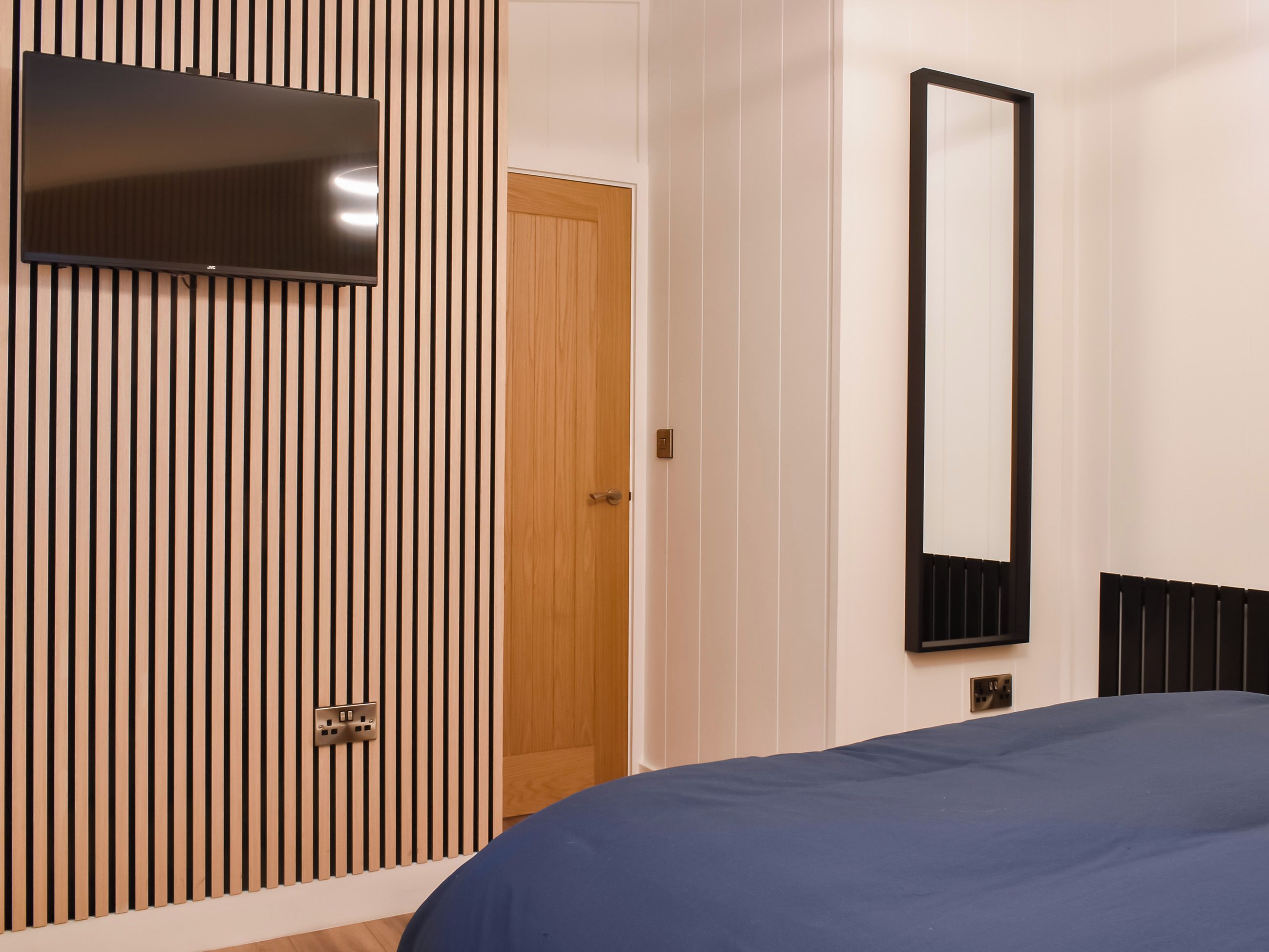 A bedroom with a bed, a TV mounted on a wall, a door, and a mirror.