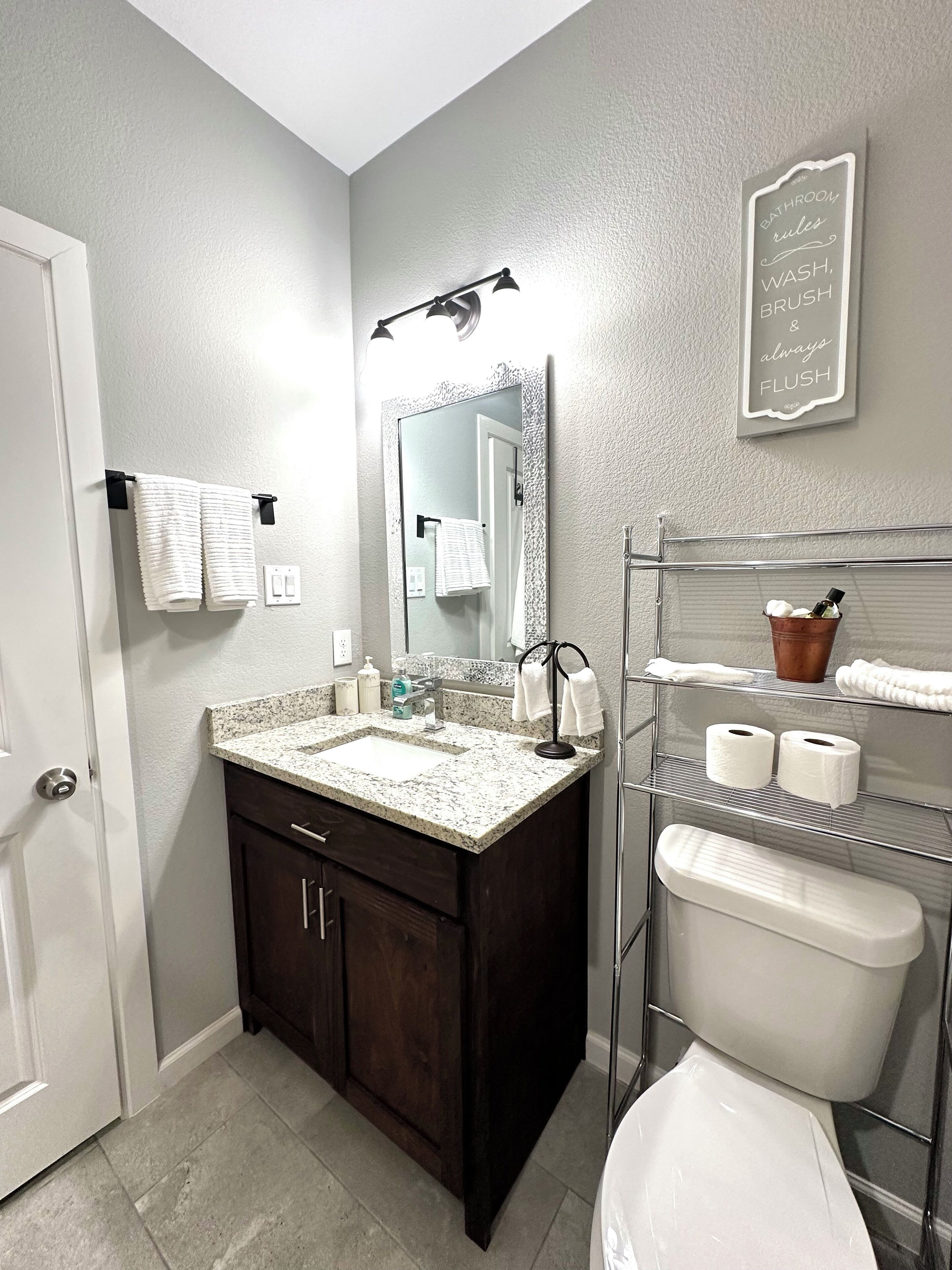 A bathroom with a dark wood vanity, a white sink, and a mirror. There is a towel rack with two towels, a shelf with toiletries, and a sign above the mirror.