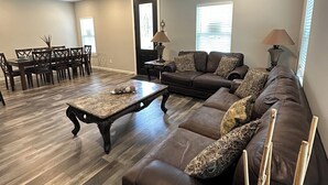 A spacious living room with a dark leather sofa, a wooden coffee table, a dining area with chairs, and a ceiling fan.