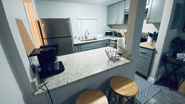Una cocina compacta con refrigerador, microondas y cafetera. Hay dos taburetes de madera y un tapete estampado.