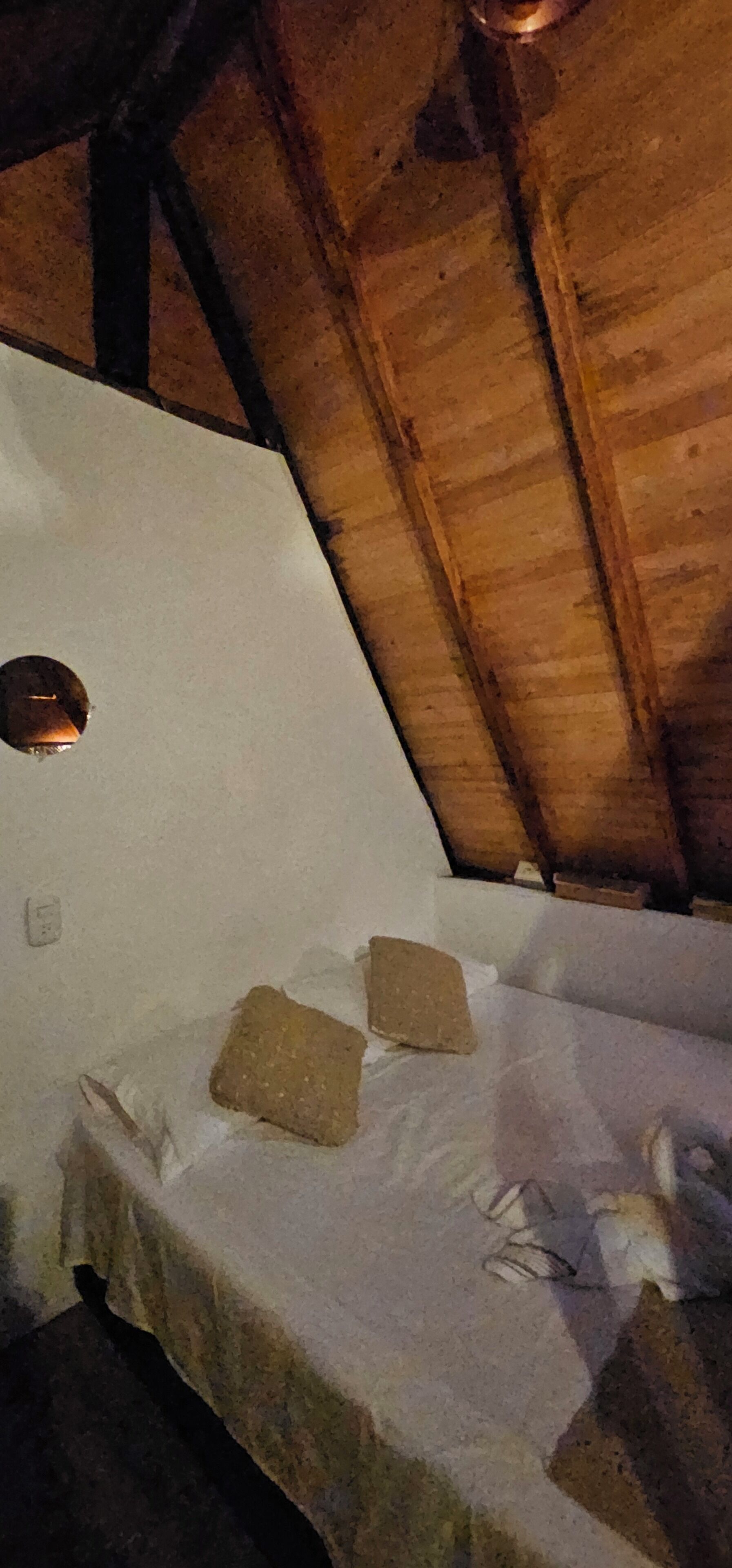 A room with a wooden ceiling, a white mattress, and two pillows on a white bedspread.