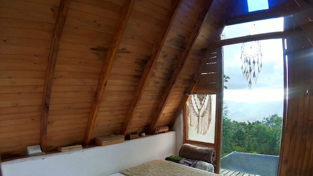 A wooden ceiling, a bed with a white sheet, a view of the outdoors through a window, and a dreamcatcher hanging on the wall. - Glamping Volver a la Tierra (Medellín)
