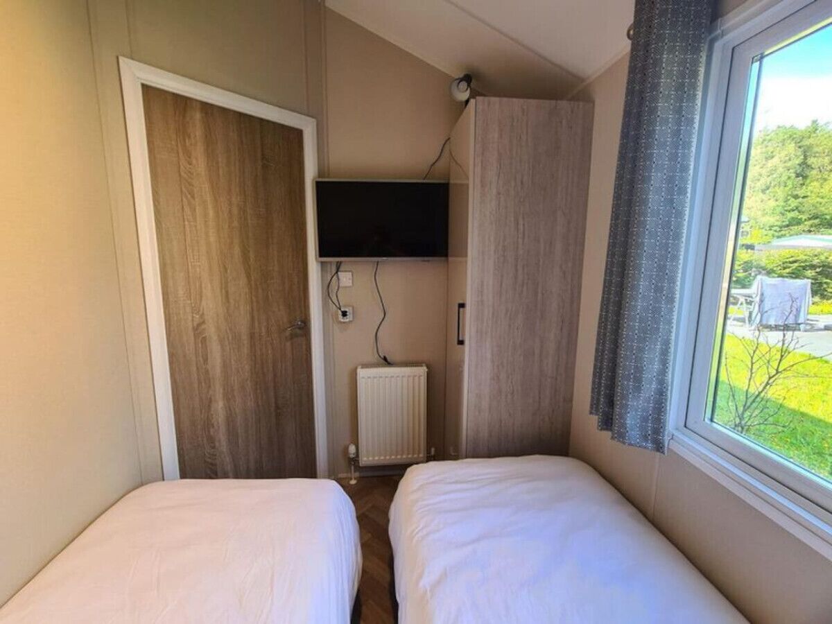 Une petite pièce avec deux lits, un téléviseur fixé au mur, un radiateur et une fenêtre avec des rideaux.