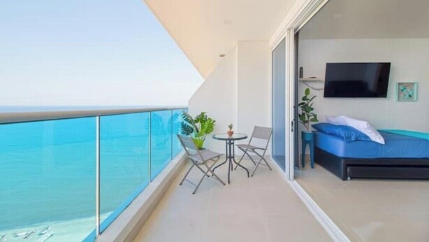 A balcony with a glass railing, a small table with chairs, a potted plant, and a bed with a blue mattress and pillows. - The Beach is Your Backyard! Ocean front, mountain views, and amazing sunset! (Santa Marta)
