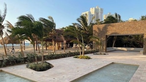 A pool area with a stone deck, palm trees, and a building with a thatched roof in the background. - The Beach is Your Backyard! Ocean front, mountain views, and amazing sunset! (Santa Marta)