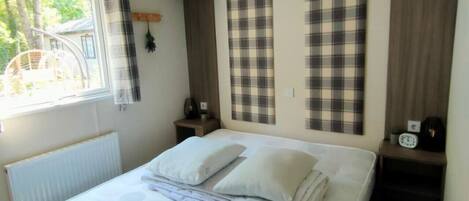 A neatly made bed with pillows, a bedside table with a clock, and plaid curtains on a window.