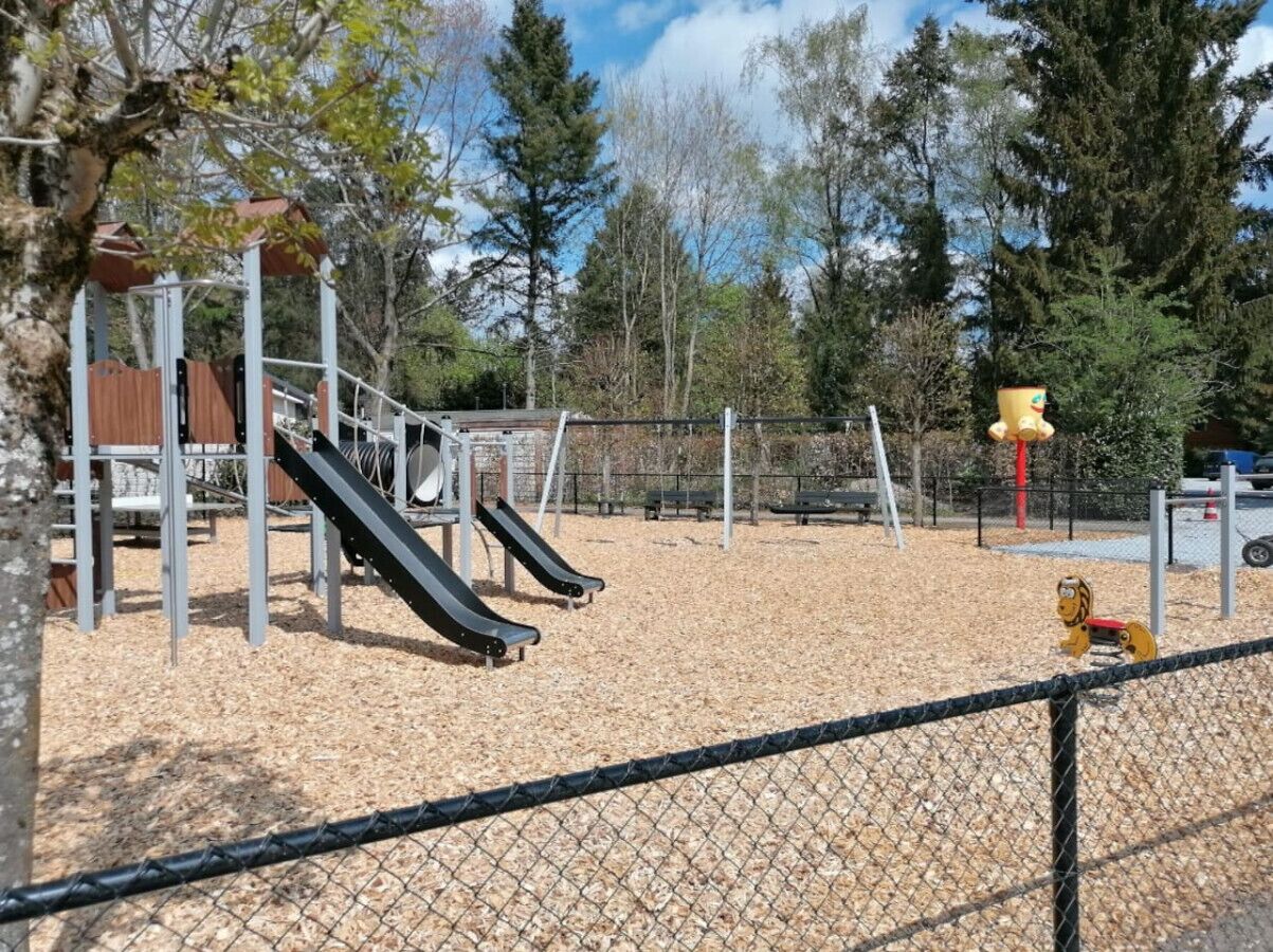 A playground with slides and a climbing structure, surrounded by a fence and trees.