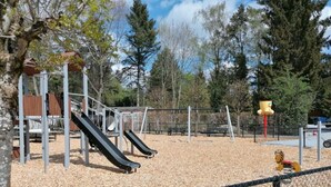 A playground with slides and a climbing structure, surrounded by a fence and trees.