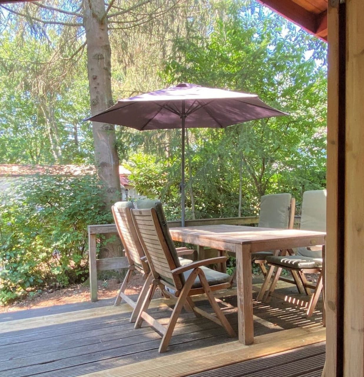 Una terraza de madera al aire libre con una mesa, sillas y una sombrilla, con vistas a una zona boscosa.