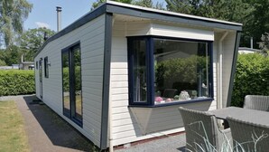 Een laag, klein gebouw met een groot raam en een deur, omringd door groen en een terras met rieten meubels.