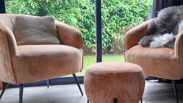 Een moderne woonkamer met twee fauteuils en een poef, een kamerkleed met een patroon, en uitzicht op groen door grote ramen.