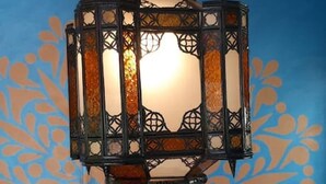 Een eetruimte met een tafel gedekt voor twee personen, een decoratieve wand met een blauw en oranje patroon en een hanglamp met een bloemendecoratie.