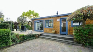 Un bâtiment en bois avec une porte et des fenêtres bleues, une terrasse pavée et un petit espace extérieur aménagé pour s’asseoir.