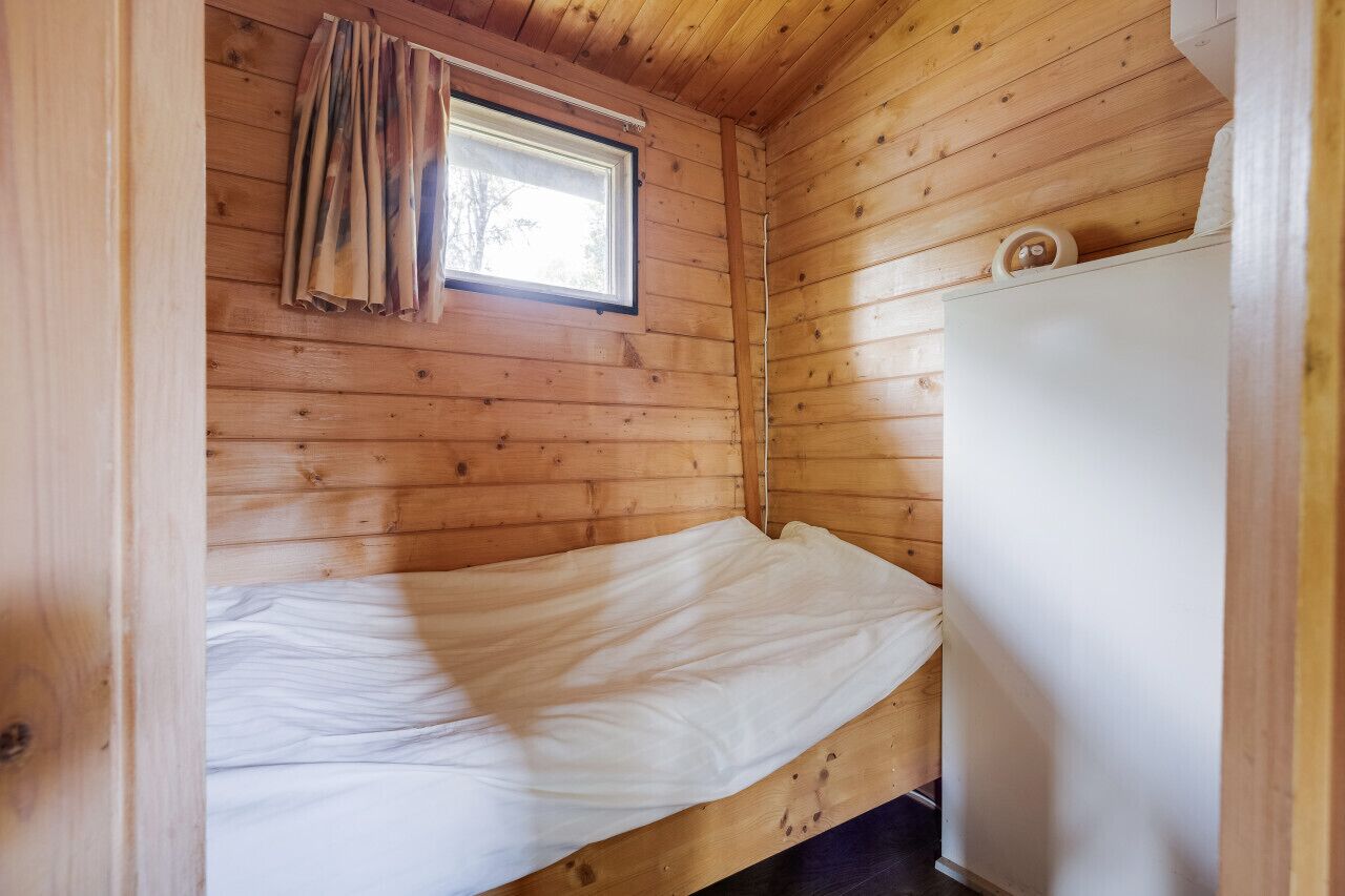 A small, single-bed cabin with wooden walls, a window with curtains, and a white cabinet.