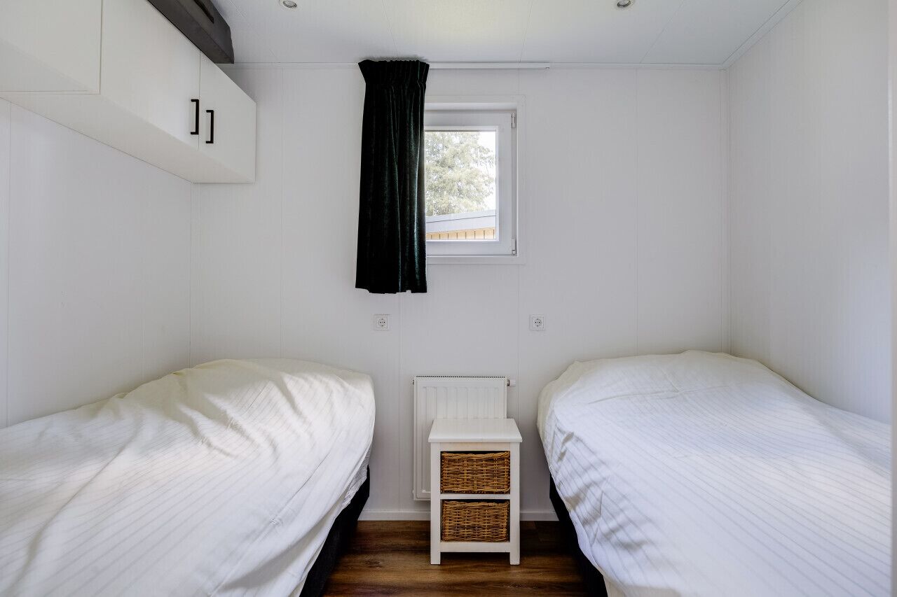 A small, clean room with two single beds, a white radiator, a small white table with baskets, and a window with a black curtain.