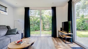 Ett modernt vardagsrum med en grå soffa, ett träbord, en tv på ett tv-bord och en skjutdörr i glas som leder ut till en trädgård.
