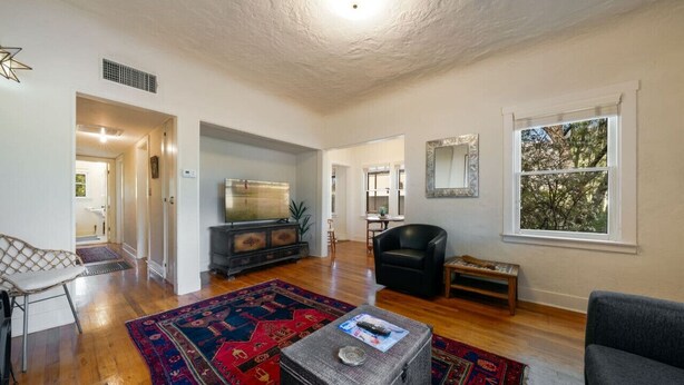 A spacious living room with hardwood floors, a large flat-screen TV, a black leather sofa, and a red patterned rug. - Charming / Outdoor Dining / 1 BR 1 BA / Sam Hughes (Tucson)