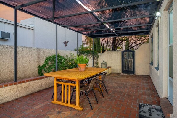 Un patio cubierto con una mesa y sillas de madera, rodeado de suelo de ladrillo y barandilla de metal.
