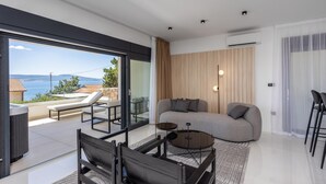 Ein modernes Wohnzimmer mit einer Couch, einem Couchtisch und einem Essbereich mit Stühlen. Ein großes Fenster bietet Blick auf das Meer und die Hügel.