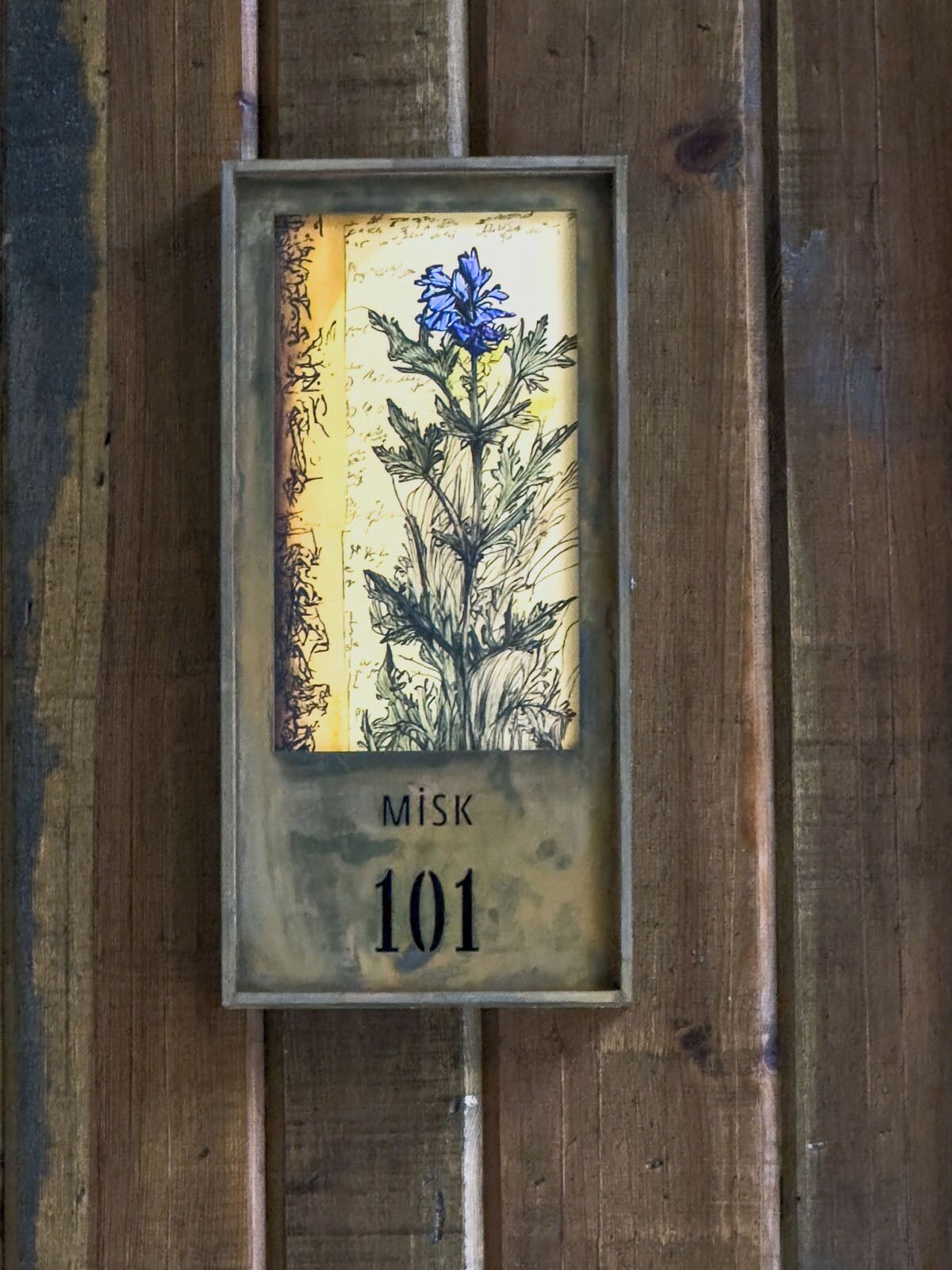 A tarot card labeled 'Misk 101' with a blue flower illustration, framed and displayed on a wooden surface.
