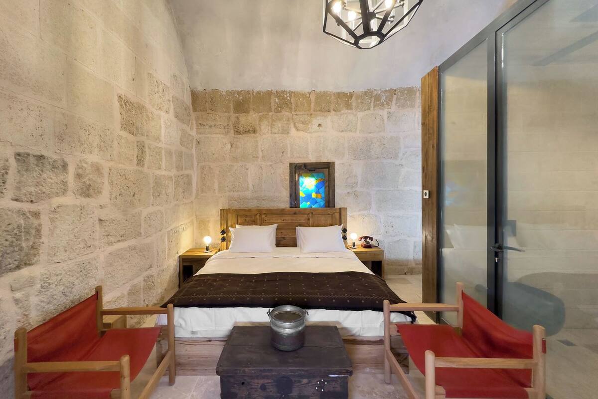 Una habitación de paredes de piedra con una cama, dos mesitas de noche, un cabecero de madera, un ventanal de vitral y una puerta de cristal.