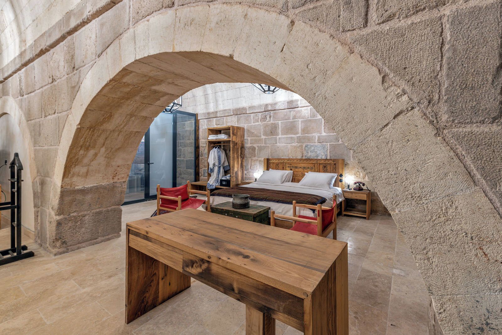 Una habitación rústica con un arco de piedra, una cama de madera, una silla roja y una mesa de madera.