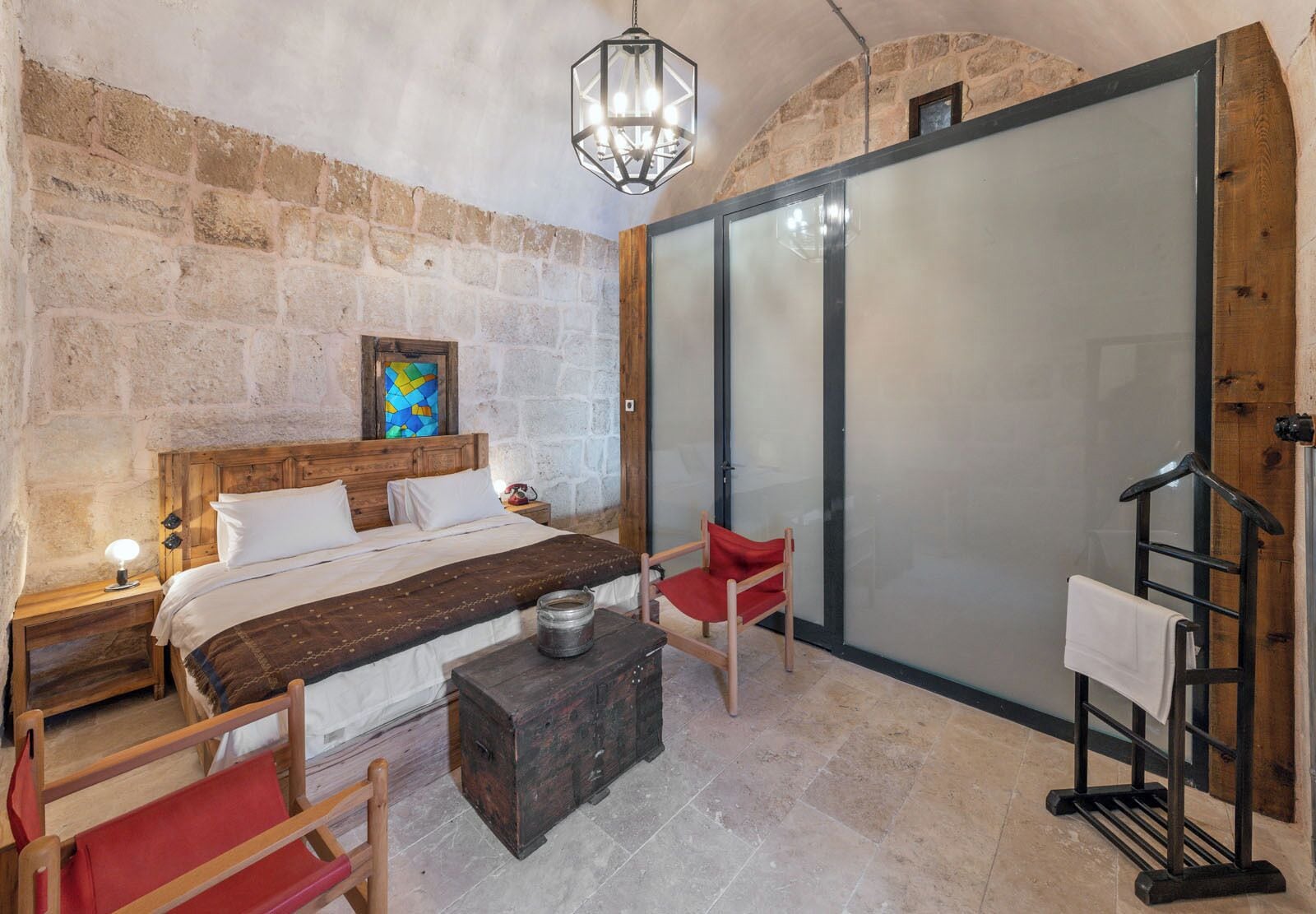 A bedroom with a stone wall, a bed with white bedding, a wooden headboard, a red chair, a small table, and a hanging light fixture.