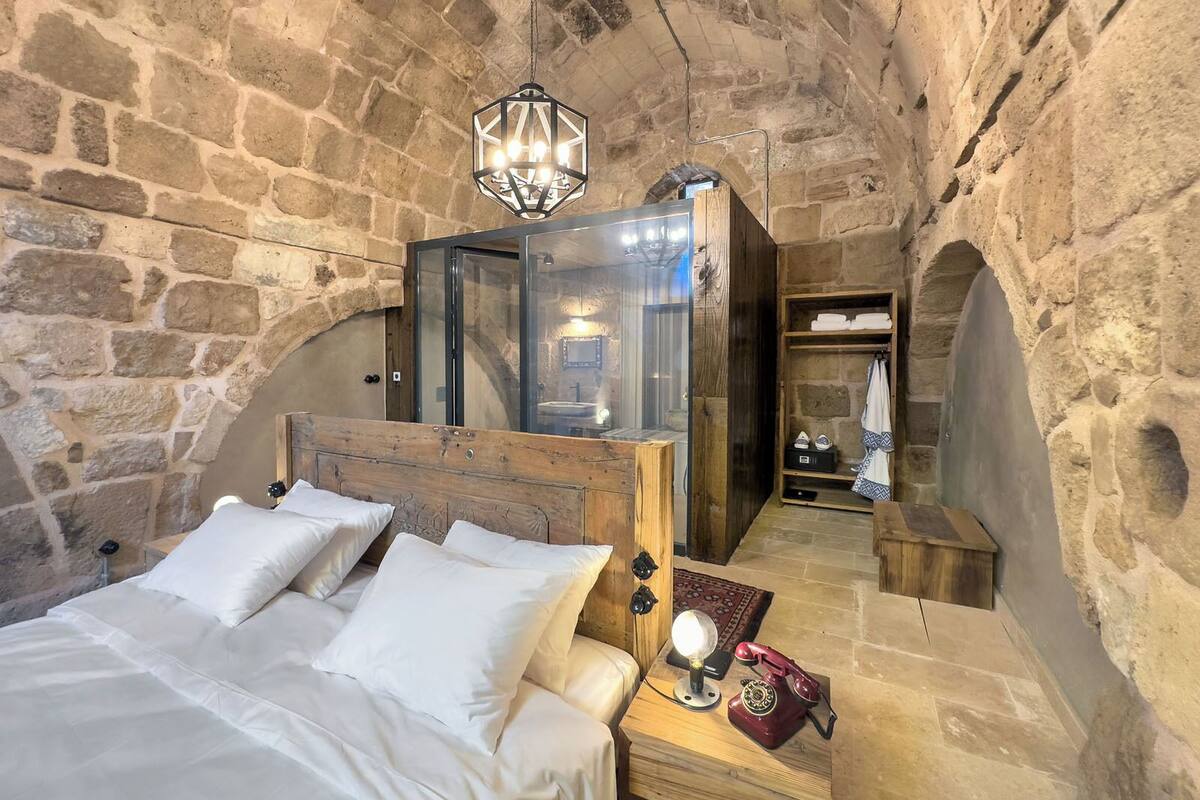 Un dormitorio con paredes de piedra, una cama de madera, un baño con cerramiento de vidrio y un teléfono antiguo en la mesita de noche.