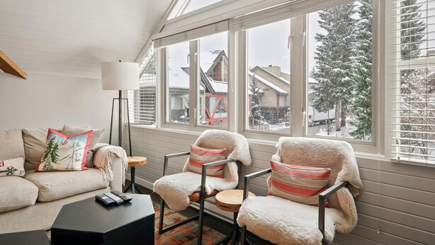 Een gezellige woonkamer met een bank, twee fauteuils, een salontafel en een vloerlamp. Grote ramen bieden uitzicht op een besneeerd landschap en gebouwen.