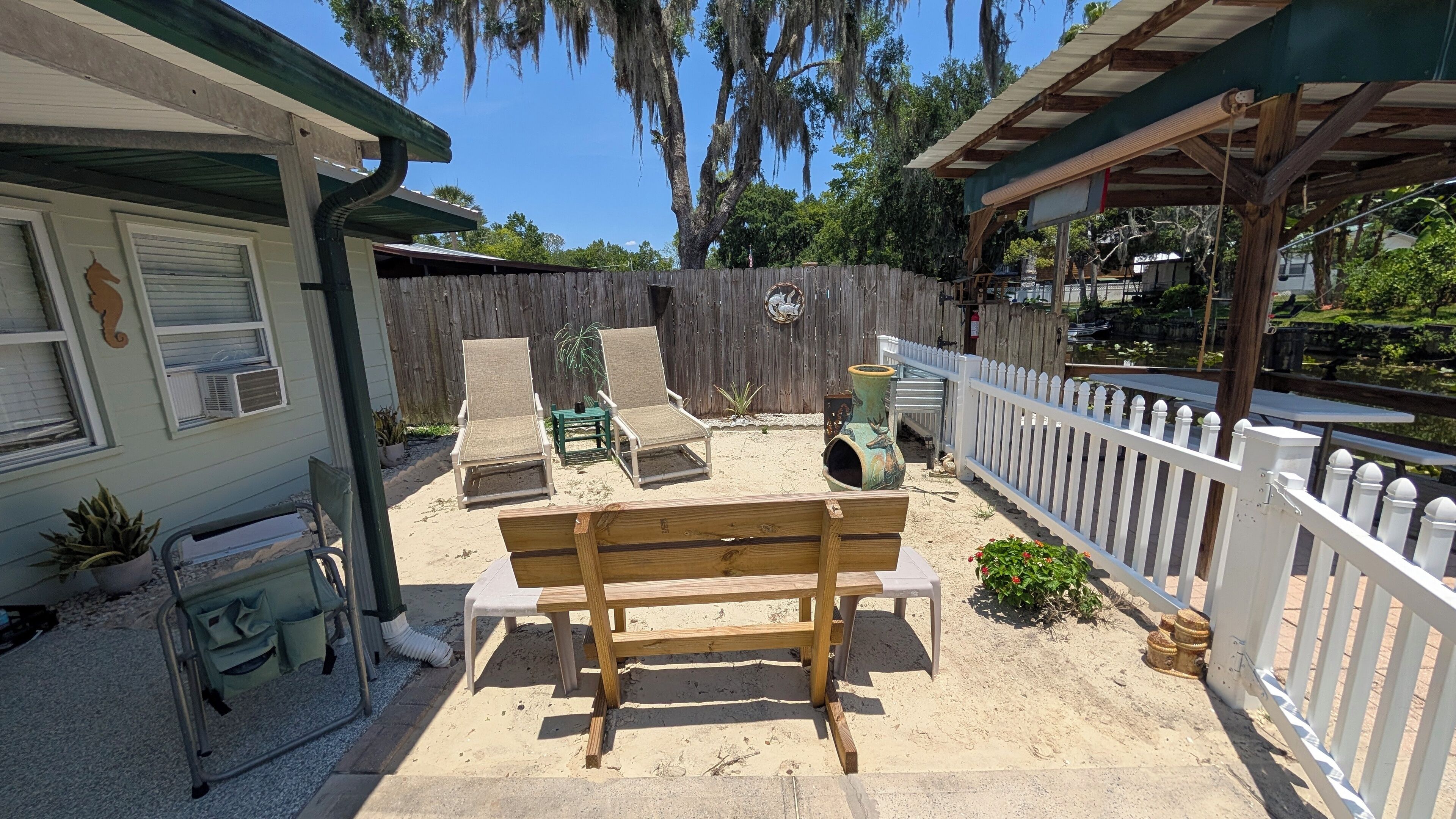 A wooden bench in a backyard with a sandy floor, a white picket fence, and a wooden deck.