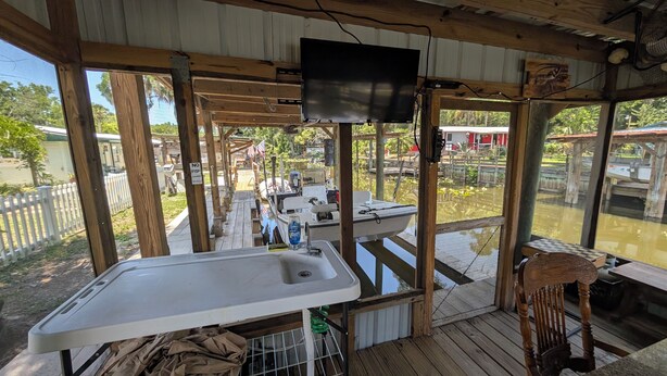 A covered outdoor area with a wooden deck, a white sink, and a television mounted on the ceiling.