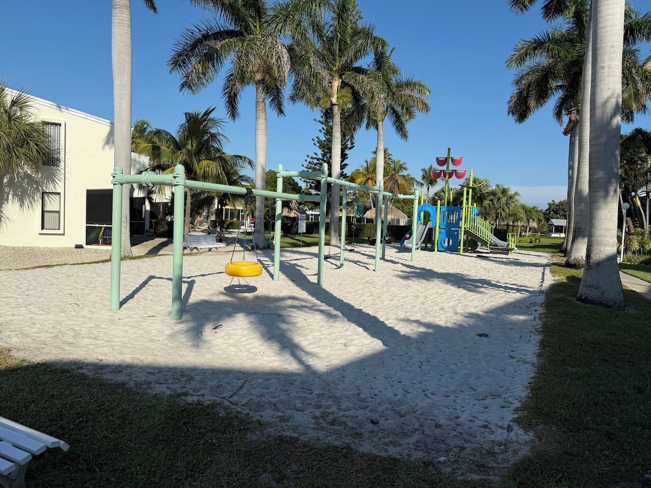 Ein Spielplatz mit Schaukeln und einer Rutsche, umgeben von Palmen und einem Gebäude.