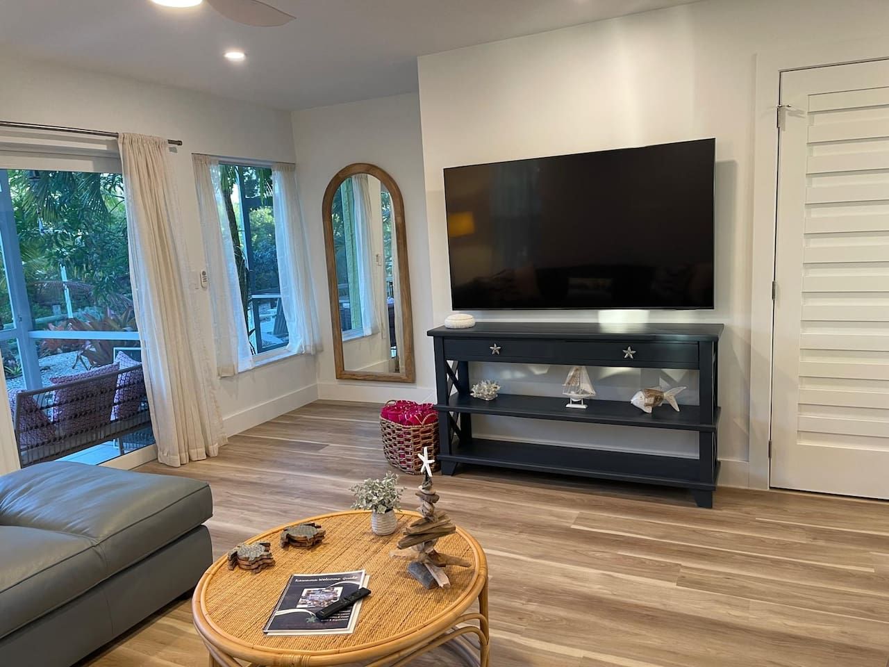 Una sala de estar moderna con un televisor de pantalla plana, un mueble de madera oscura para entretenimiento, una mesa de centro de mimbre con objetos decorativos, un sofá gris y ventanas grandes con cortinas.