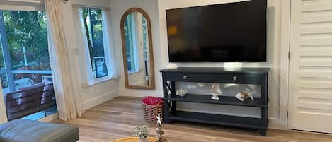 Una sala de estar moderna con un televisor de pantalla plana, un mueble de madera oscura para entretenimiento, una mesa de centro de mimbre con objetos decorativos, un sofá gris y ventanas grandes con cortinas.